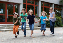 Fünf fröhliche Kinder rennen lachend über einen Schulhof. Sie tragen legere Kleidung und scheinen Spaß an der Bewegung zu haben. Im Hintergrund ist ein modernes Schulgebäude mit großen Fenstern zu sehen. Das Bild vermittelt ein Gefühl von unbeschwerter Kindheit und Bewegung und betont die Bedeutung von sicheren Spiel- und Bewegungsräumen im Rahmen der Unfallverhütung und Prävention für Kinder, ein Anliegen der Unfallkasse NRW.