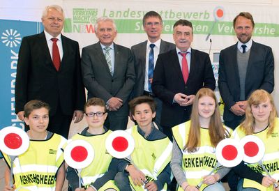 Eine Gruppe von neun Personen posiert bei einer Preisverleihung. Im Hintergrund ist das Logo der Unfallkasse NRW sichtbar. In der oberen Reihe stehen vier Männer in Anzügen und einer in einer Freizeitjacke. In der unteren Reihe knien fünf Jugendliche in gelben Sicherheitswesten mit der Aufschrift 'Verkehrshelfer'. Sie halten rote und weiße Schilder. Der Kontext ist die Förderung von Sicherheit und Prävention im Schulwegverkehr.