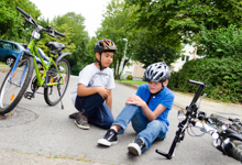 Zwei Kinder mit Fahrradhelmen sitzen auf einer Straße neben einem umgekippten Fahrrad. Einer der Jungen hält sein Knie, als wäre er gestürzt. Das Bild vermittelt die Bedeutung von Schutzmaßnahmen und Vorsicht beim Fahrradfahren. Im Hintergrund sind Bäume und Wohnhäuser zu sehen, die ein Wohngebiet andeuten. Das Bild könnte auf die Notwendigkeit der Unfallprävention hinweisen, insbesondere im Hinblick auf Kindersicherheit im Straßenverkehr.