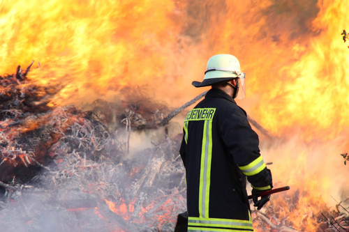 Ein Feuerwehrmann steht mit Schutzkleidung und Helm vor einer großen, lodernden Flammenwand. Er hält ein Werkzeug in der Hand und arbeitet inmitten von Rauch und brennendem Unterholz. Die Szene verdeutlicht die Herausforderungen und Risiken, die Einsatzkräfte im Rahmen des Arbeitsschutzes und der Prävention bewältigen.