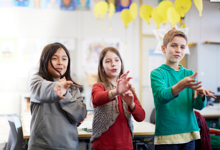 Drei Kinder stehen in einem Klassenzimmer und üben Gesten, möglicherweise im Rahmen eines Präventionsprogramms. Die Gruppe wirkt konzentriert und engagiert. Im Hintergrund sind unscharf Schulposter und gelbe Dekorationen zu erkennen. Das Bild betont die Bedeutung von Bildung und Prävention in der Arbeit der Unfallkasse NRW.