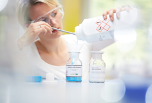 Eine Frau trägt eine Schutzbrille und füllt aus einem großen Plastikbehälter eine blaue Flüssigkeit in ein Glasgefäß. Ein weiteres Glasgefäß mit einer anderen Substanz steht daneben. Die Umgebung sieht wie ein Labor aus. Diese Szene betont die Wichtigkeit von Schutzmaßnahmen und Prävention bei der Handhabung von Chemikalien, relevant für Arbeitsschutz und Prävention durch die Unfallkasse NRW.