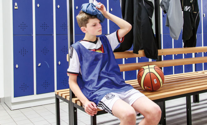 Ein junger Schüler sitzt auf einer Holzbank in einem Umkleideraum mit blauen Spinden. Er trägt ein blaues Trainingshemd über einem weißen Trikot und hält einen Eisbeutel an seinen Kopf. Vor ihm liegt ein Basketball auf der Bank. Die Szene deutet auf Erste-Hilfe-Maßnahmen bei einer Sportverletzung hin, was die Bedeutung von Sicherheitsmaßnahmen und Verletzungsprävention im Schulsport unterstreicht.