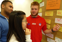 Drei junge Erwachsene stehen vor einer Pinnwand, die mit farbigen Notizzetteln bedeckt ist. Eine Person im roten Hoodie erklärt etwas und zeigt auf die Notizen, die möglicherweise Themen zu Arbeitsschutz und Prävention betreffen. Eine andere Person im blauen Pullover und eine dritte mit Brille hören aufmerksam zu. Die Szene erfolgt in einem Kontext, der auf eine Bildungs- oder Trainingssituation für die Unfallkasse NRW hindeutet.