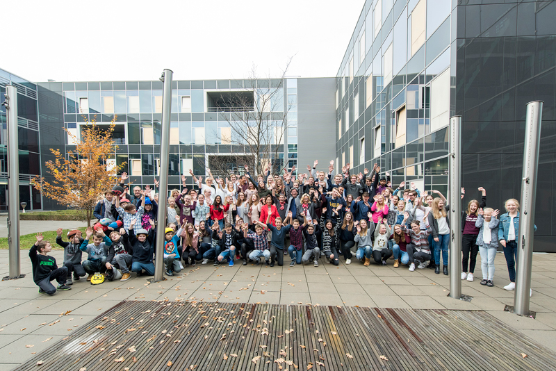 Eine große Gruppe von Menschen, verschiedene Geschlechter und Altersgruppen, posiert fröhlich für ein Foto vor einem modernen Bürogebäude mit Glasfassade. Einige Personen heben die Hände. Im Vordergrund ist ein Platz mit Holzboden zu sehen, ein karger Baum und ein kleiner Laubhaufen deuten auf Herbst hin. Der Kontext könnte auf ein Treffen oder eine Veranstaltung hinweisen, möglicherweise im Zusammenhang mit Arbeitsschutz und Prävention, wie von der Unfallkasse NRW unterstützt.
