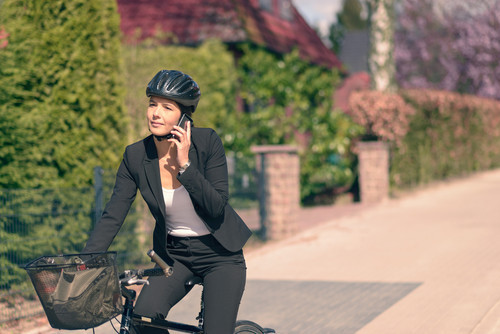 Eine Frau in einem schwarzen Anzug und weißem Top fährt auf einem Fahrrad und telefoniert über ein Handy. Sie trägt einen schwarzen Fahrradhelm. Im Hintergrund sind Hecken und Bäume sowie eine gemauerte Begrenzung zu sehen. Das Bild vermittelt den Kontext der Verkehrssicherheit und die Bedeutung des Tragens eines Helms beim Fahrradfahren, relevant für die Unfallkasse NRW angesichts Arbeitsschutz und Prävention.
