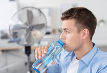 Ein junger Mann in einem hellblauen Hemd sitzt in einem Büro und trinkt aus einer Wasserflasche. Im Hintergrund steht ein großer Ventilator. Das Bild vermittelt wichtige Aspekte des Arbeitsschutzes, wie regelmäßige Hydration und Kühlung, um Hitzestress am Arbeitsplatz zu vermeiden. Dies ist besonders relevant für die Präventionsmaßnahmen der Unfallkasse NRW.