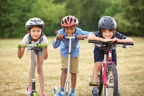 Drei Kinder stehen nebeneinander auf einer Wiese. Zwei Kinder haben Roller, eines hat ein Fahrrad. Alle tragen Helme. Die Szene vermittelt Sicherheit und Prävention im Kontext von Freizeitaktivitäten für Kinder und Arbeitsschutz. Im Hintergrund ist verschwommenes, grünes Laubwerk zu sehen, was auf einen Park hindeuten könnte.