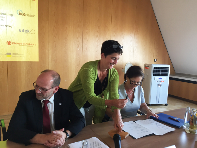 Drei Personen sitzen an einem rechteckigen Tisch in einem Konferenzraum mit Holzverkleidung. Zwei Frauen und ein Mann tragen Business-Kleidung, sie beschäftigen sich mit Papierdokumenten. Die Frau in der Mitte steht und erklärt etwas den anderen. Auf dem Tisch sind Mikrofone und Wasserflaschen platziert. Im Hintergrund sind Logos und ein Klimagerät zu sehen. Kontext: Unfallkasse NRW, Arbeitsschutz, Prävention.