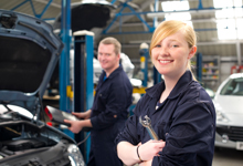 In einer Autowerkstatt stehen zwei Menschen in Arbeitskleidung. Eine junge Frau im Vordergrund hält einen Schraubenschlüssel und lächelt, während ein Mann im Hintergrund mit einem Diagnosewerkzeug an einem Fahrzeug arbeitet. Der Fokus liegt auf der Bedeutung von Arbeitsschutz und Prävention in der Kfz-Branche, unterstützt von der Unfallkasse NRW.
