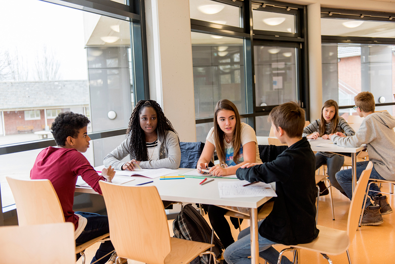 Sechs Jugendliche sitzen an Tischen in einem hellen, modernen Raum mit großen Fenstern. Sie diskutieren angeregt und arbeiten mit Papier und Stiften. Der Raum hat helle Wände und eine angenehme Atmosphäre. Die Szene symbolisiert Teamarbeit und Kommunikation, wichtige Aspekte im Bereich Arbeitsschutz und Prävention.