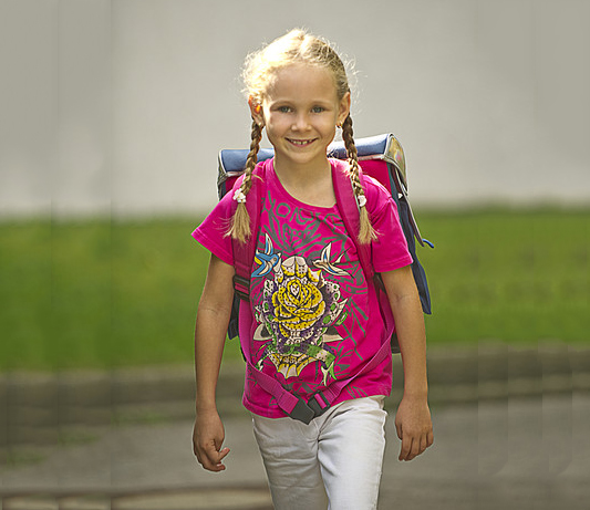 Ein junges Mädchen mit blonden Zöpfen und einem pinkfarbenen T-Shirt trägt einen Schulranzen und lächelt während sie im Freien geht. Im Hintergrund sind Gras und ein unscharfer Gebäudeteil sichtbar. Das Bild vermittelt ein Gefühl von Schulbeginn und Sicherheit auf dem Weg zur Schule, was im Kontext der Unfallkasse NRW für die Bedeutung von Prävention und Arbeitsschutz steht.