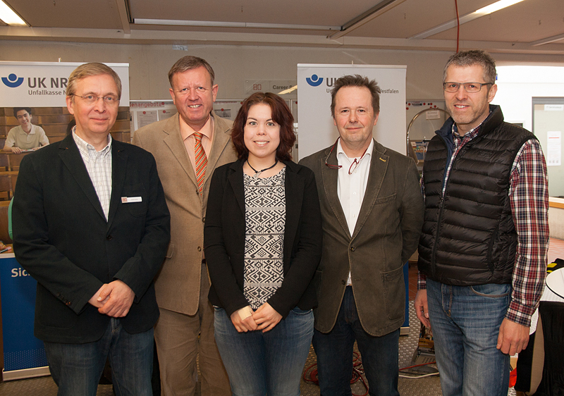 Fünf Personen stehen vor Bannern der Unfallkasse NRW in einem Raum. Die Banner im Hintergrund tragen das Logo der UK NRW und vermitteln den Fokus auf Sicherheit und Arbeitsschutz. Der Raum scheint für eine Veranstaltung oder Präsentation vorbereitet zu sein, mit zusätzlicher Technik und Informationsmaterialien. Dieses Setting deutet auf eine Besprechung oder ein Seminar zur Prävention von Arbeitsunfällen hin.