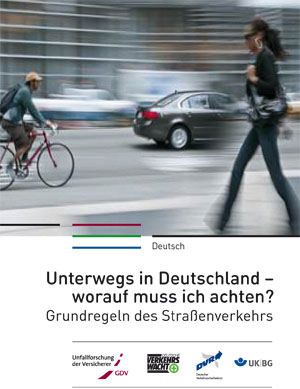 Das Bild zeigt ein Plakat mit dem Titel 'Unterwegs in Deutschland – worauf muss ich achten? Grundregeln des Straßenverkehrs'. Im Hintergrund sind verschwommene Personen und Verkehrsfahrzeuge auf einer Straße erkennbar, was die Bewegung und den Straßenverkehr symbolisiert. Das Plakat enthält Logos der Unfallforschung der Versicherer, Verkehrswacht, DVR und UK/IBG, was auf die Beteiligung dieser Organisationen an der Verkehrssicherheitskampagne hinweist.