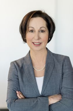 Das Bild zeigt eine lächelnde Frau mittleren Alters in einem grauen Blazer mit einem weißen Oberteil darunter. Die Frau hat dunkles, kinnlanges Haar und trägt eine Perlenkette. Sie steht vor einem neutralen, weißen Hintergrund und hat die Arme vor dem Körper verschränkt. Möglicher Kontext zu Arbeitsschutz und Prävention für die Unfallkasse NRW.