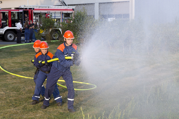 Drei Jugendliche in blauer Feuerwehrschutzkleidung und orangefarbenen Helmen stehen auf einer Rasenfläche und halten gemeinsam einen Feuerwehrschlauch, aus dem ein starker Wasserstrahl kommt. Im Hintergrund befindet sich ein rotes Feuerwehrfahrzeug und mehrere Personen, die die Übung beobachten. Die Szene vermittelt ein Gefühl von Teamarbeit und Ausbildung im Rahmen von Arbeitsschutz- und Präventionsmaßnahmen durch die Unfallkasse NRW.