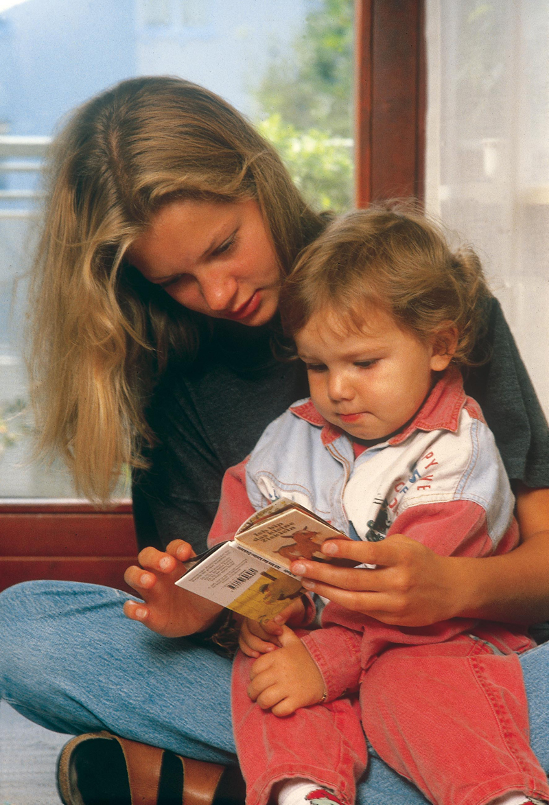 Eine Erzieherin sitzt mit einem Kleinkind auf dem Boden und liest ein Kinderbuch. Beide sind konzentriert und interagieren intensiv miteinander. Die Szene spielt in einem geschützten, sicher wirkenden Innenraum mit natürlichem Licht, was auf einen sorgsamen Umgang mit der Umgebung hinweist. Der Fokus liegt auf Bildungsförderung und der Sicherheit in der Kinderbetreuung, passend zu Themen der Unfallkasse NRW.