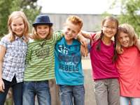 Ein fröhliches Bild von fünf Kindern, die draußen stehen und sich gegenseitig umarmen. Sie lächeln in die Kamera. Die Szene vermittelt Gemeinschaft und positive Interaktion, im Einklang mit Themen der Unfallkasse NRW wie Arbeitsschutz und Prävention, indem sie betont, wie wichtig es ist, schon in jungen Jahren aufeinander zu achten.