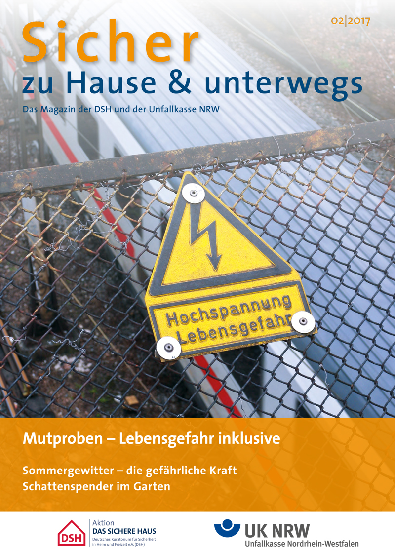 Titelbild der Zeitschrift 'Sicher zu Hause & unterwegs' der DSH und Unfallkasse NRW. Im Vordergrund ist ein gelbes Warnschild mit der Aufschrift 'Hochspannung Lebensgefahr' an einem Maschendrahtzaun zu sehen, der ein Bahngleisbereich abgrenzt. Der obere Bereich des Covers ist blau mit weißer und oranger Schrift. Der orangefarbene untere Teil enthält Artikelvorschläge: 'Mutproben – Lebensgefahr inklusive', 'Sommergewitter – die gefährliche Kraft', und 'Schattenspender im Garten'. Das Logo der Unfallkasse Nordrhein-Westfalen befindet sich unten rechts.
