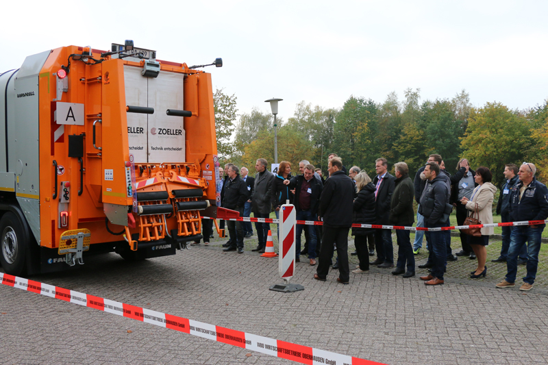 Eine Gruppe von Menschen steht um ein orangefarbenes Müllfahrzeug herum, das für Sicherheitsdemonstrationen präsentiert wird. Absperrbänder trennen die Zuschauer von dem Fahrzeug. Im Hintergrund sind Bäume mit Herbstlaub zu sehen. Die Szene findet auf einer gepflasterten Fläche im Freien statt, was auf eine Veranstaltung im Bereich Arbeitsschutz und Prävention hindeutet, organisiert von der Unfallkasse NRW.