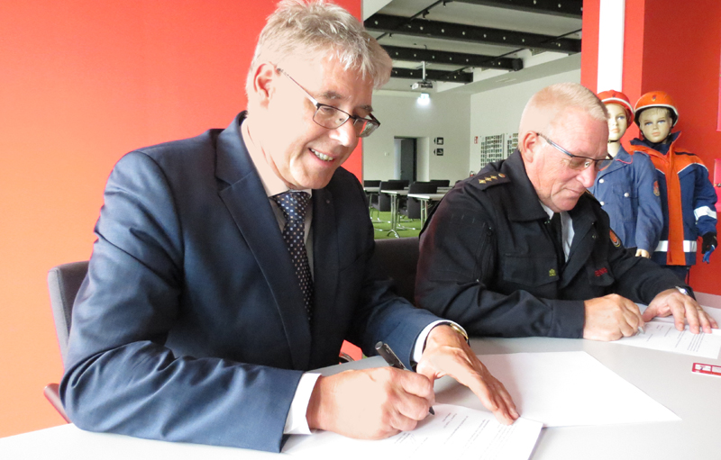 Zwei Männer, einer in Anzug, der andere in Feuerwehruniform, sitzen an einem Tisch in einem Büro mit roten Wänden. Sie unterzeichnen Dokumente. Im Hintergrund sind Schaufensterpuppen in Feuerwehrkleidung. Der Kontext deutet auf eine Maßnahme zur Arbeitssicherheit und Prävention im Zusammenhang mit der Unfallkasse NRW hin.