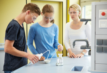 Drei junge Personen stehen in einem Labor. Zwei von ihnen mischen Substanzen in einem Becherglas, während die dritte Person zusieht. Alle tragen Schutzbrillen, um ihre Augen zu schützen, was auf die Wichtigkeit von Arbeitsschutz und Prävention im Laboralltag hinweist. Im Hintergrund sind Laborgeräte und helle Wände zu sehen.