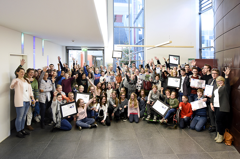 Eine Gruppe von Menschen, bestehend aus unterschiedlichen Altersgruppen, steht in einem großen, hellen Raum und posiert für ein Foto. Einige halten Urkunden in die Höhe, während andere die Arme jubelnd nach oben strecken. Der Raum hat große Fenster im Hintergrund, die viel Licht hereinlassen. Dies könnte eine Veranstaltung der Unfallkasse NRW sein, die den Fokus auf Arbeitsschutz und Prävention legt.