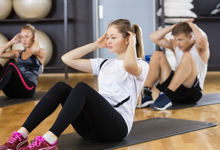 Drei junge Erwachsene führen gemeinsam Bauchübungen auf Matten in einem Fitnessstudio durch. Im Hintergrund sind große Gymnastikbälle und gestapelte Handtücher zu sehen. Die Szene vermittelt den Aspekt der Gesundheitsförderung und Prävention durch Fitnessaktivitäten, was im Kontext der Unfallkasse NRW zur Förderung von Arbeitsschutz und Prävention passt.