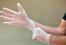 Eine Person streift sich einen weißen Einweghandschuh über die Hand. Der Fokus liegt auf dem Schutz der Hände, was für den Arbeitsschutz und die Prävention von großer Bedeutung ist. Die Szene betont die Wichtigkeit von persönlicher Schutzausrüstung am Arbeitsplatz, um Verletzungen und Kontaminationen zu vermeiden.