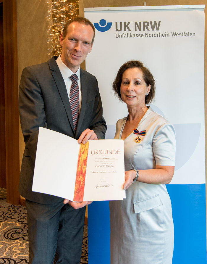 Zwei Personen präsentieren eine Urkunde vor einem UK NRW Banner.