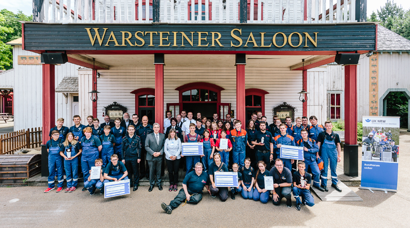 Eine große Gruppe von Menschen, darunter viele in Arbeitskleidung, steht vor dem Eingang des Warsteiner Saloons. Sie halten verschiedene Auszeichnungen und Schilder, die auf die Unfallkasse NRW hinweisen. Das Gebäude im Hintergrund hat weiße Holzverkleidung mit roten Elementen. Die Szene könnte mit einem Event im Zusammenhang mit Arbeitsschutz und Prävention in Verbindung stehen.