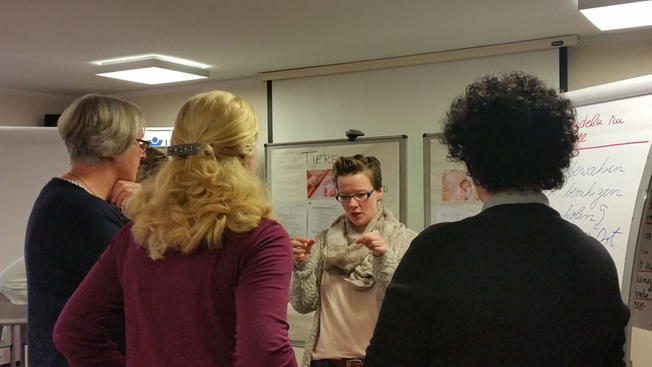 Vier Personen stehen vor einem Whiteboard und diskutieren. Eine Frau in der Mitte spricht gestikulierend, während die anderen zuhören. Auf dem Whiteboard sind grafische Darstellungen und Notizen sichtbar. Die Szene findet in einem Konferenzraum statt und thematisiert wahrscheinlich Arbeitsschutz oder Präventionsmaßnahmen im Kontext der Unfallkasse NRW.