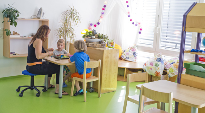 Eine Erzieherin sitzt mit zwei kleinen Kindern in einem freundlich gestalteten Kindergartenraum an einem Tisch. Der Boden ist grün, Wände sind hell, und es gibt Regale mit Pflanzen und Spielzeug. Die Kinder und die Erzieherin sind in ein Brettspiel vertieft. Der Raum ist mit hellen Möbeln und farbenfrohen Kissen eingerichtet. Das Bild vermittelt eine sichere und anregende Umgebung, die für die Arbeitsschutzkonzepte der Unfallkasse NRW im Bereich Prävention und Sicherheit in Bildungseinrichtungen steht.