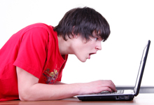 Ein Jugendlicher mit braunen Haaren trägt ein rotes T-Shirt und schaut überrascht auf einen Laptop-Bildschirm. Das Bild könnte auf Themen wie sichere Computernutzung, Ergonomie am Arbeitsplatz oder digitale Ablenkung hinweisen. Der sichere und verantwortungsvolle Umgang mit digitalen Medien ist besonders wichtig, um gesundheitlichen Risiken im Arbeits- und Bildungsumfeld vorzubeugen.