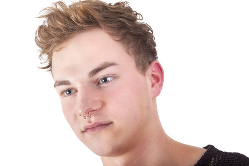 Das Bild zeigt das Gesicht eines jungen Mannes mit leicht gelocktem, hellbraunem Haar und einem Nasenpiercing. Er trägt einen dunkel gestrickten Pullover und blickt leicht zur Seite. Der Hintergrund ist weiß. Im Kontext der Unfallkasse NRW könnte das Bild für Themen des persönlichen Schutzes und der individuellen Ausdrucksweise am Arbeitsplatz stehen.