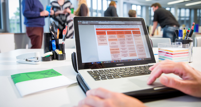 In einem modernen Büro arbeitet eine Person an einem Laptop. Der Bildschirm zeigt eine Tabelle mit rotem und weißem Layout. Im Hintergrund sind andere Personen unscharf zu sehen, die miteinander sprechen und stehen. Auf dem Tisch liegen Büroartikel wie Stifte, Notizblöcke und ein Locher. Diese Umgebung könnte auf Schulungen oder Besprechungen im Kontext von Arbeitsschutz und Prävention hinweisen, die für die Unfallkasse NRW relevant sind.