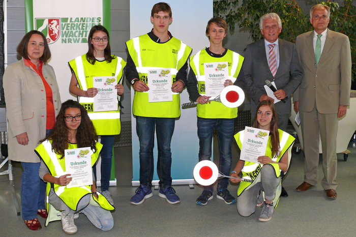 Sechs Personen stehen nebeneinander in einem Raum. Vier Jugendliche in gelben Warnwesten halten Urkunden in den Händen. Zwei von ihnen tragen zusätzlich Signaltafeln. Links und rechts stehen zwei Erwachsene in formeller Kleidung, die das Bild formal und ernst erscheinen lassen. Im Hintergrund sind ein Banner mit dem Logo der Landesverkehrswacht NRW und der Schriftzug erkennbar. Das Bild hat Bezüge zum Thema Arbeitsschutz und Prävention.