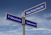 Ein Straßenschild mit drei blauen Schildern zeigt in unterschiedliche Richtungen und trägt die Aufschriften 'Frühschicht', 'Nachtschicht' und 'Spätschicht'. Der Hintergrund ist ein bewölkter Himmel. Das Bild verweist auf die Themen Arbeitsschutz und Prävention im Kontext von Schichtarbeit, relevant für die Unfallkasse NRW.