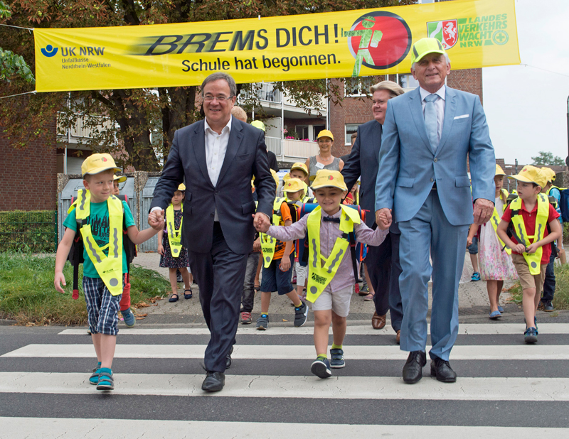Zwei Erwachsene in Anzügen begleiten zwei Kinder, die leuchtend gelbe Westen und Mützen tragen, über einen Fußgängerüberweg. Im Hintergrund folgen weitere Kinder mit identischer Sicherheitskleidung. Über ihnen ist ein gelbes Banner mit der Aufschrift 'Brems dich! Schule hat begonnen.' sichtbar. Links oben das Logo der Unfallkasse NRW, rechts das der Landesverkehrswacht. Die Szene suggeriert einen Schulbeginn und betont die Wichtigkeit von Vorsichtsmaßnahmen im Straßenverkehr.