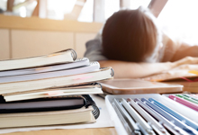 Einheiten von Ordnern und Dokumenten liegen gestapelt auf einem Tisch, während eine Person mit dem Kopf auf dem Tisch in der Nähe liegt. Der helle Hintergrund deutet auf Tageslicht und ein Büro oder einen Arbeitsplatz hin. Das Bild vermittelt das Thema Überlastung und Ermüdung am Arbeitsplatz, ein wichtiger Aspekt im Kontext von Arbeitsschutz und Prävention, relevant für die Unfallkasse NRW.