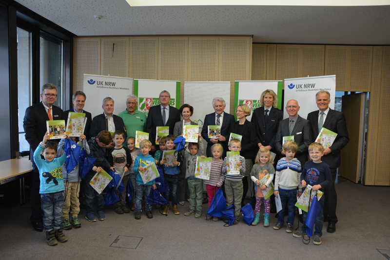 Eine Gruppe von Erwachsenen und kleinen Kindern posiert für ein Foto. Die Personen halten Broschüren und Geschenktaschen in Händen. Im Hintergrund sind zwei Roll-Up Banner mit dem Logo der Unfallkasse NRW sichtbar. Kinder tragen bunte Kleidung und stehen in einer Reihe vor den Erwachsenen. Die Veranstaltung scheint im Zusammenhang mit Arbeitsschutz und Prävention von der Unfallkasse NRW zu stehen.