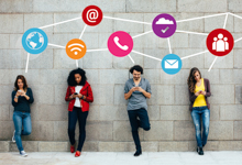 Vier junge Erwachsene stehen an einer grauen Wand und sind vertieft in ihre Smartphones. Über ihnen sind Linien mit digitalen Netzwerk- und Kommunikationsicons wie Globus, E-Mail und Telefon angebracht. Diese Darstellung symbolisiert digitale Vernetzung und Kommunikation im Alltag. Kontext: Förderung von Arbeitssicherheit und Präventionsmaßnahmen bei Nutzung digitaler Technologien.