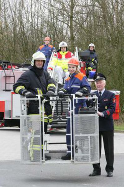 Eine Gruppe von Feuerwehrleuten in voller Schutzbekleidung steht vor einem Leiterfahrzeug. Zwei Feuerwehrleute befinden sich im Rettungskorb, der vom Fahrzeug ausgefahren ist. Im Hintergrund stehen weitere Feuerwehrkräfte am Fahrzeug. Der Einsatz zeigt die Bedeutung von Sicherheit und Arbeitsschutz bei der Feuerwehr. Die Szene findet im Freien statt, umgeben von kahlen Bäumen, was auf eine Übung im Winter oder frühen Frühling hindeutet. Diese Übung steht im Kontext der Unfallkasse NRW und betont die Präventionsmaßnahmen bei der Feuerwehr.