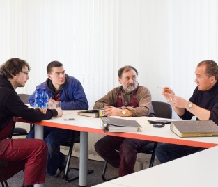 Vier Männer sitzen an einem Tisch in einem Konferenzraum. Zwei Männer tragen blaue Overalls, die anderen Freizeitkleidung. Auf dem Tisch liegen Ordner, ein Notizbuch und Handys. Einer der Männer gestikuliert, während er spricht. Der Raum hat weiße Wände und vertikale Jalousien. Der Kontext deutet auf ein Treffen zur Besprechung von Arbeitsschutzmaßnahmen oder Prävention hin.