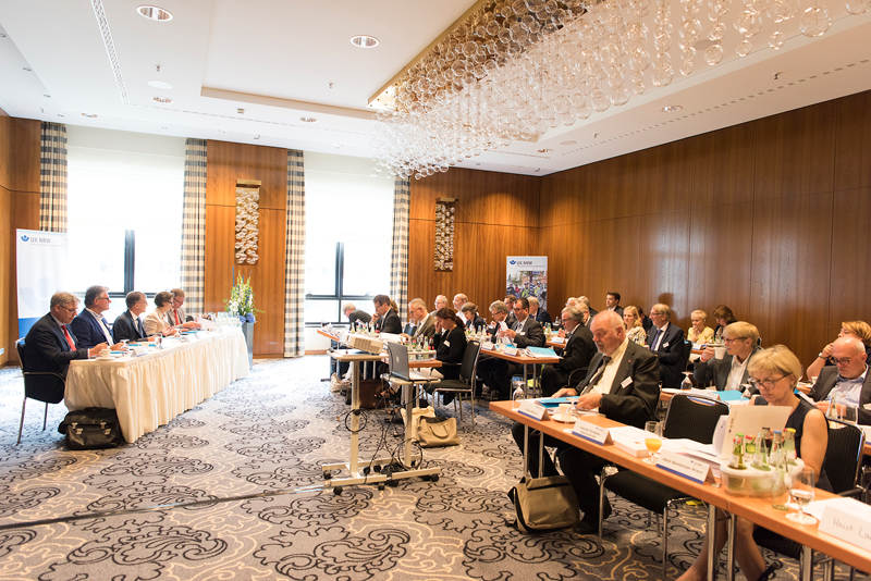 Ein Konferenzraum mit mehreren Teilnehmern, die an zwei Tischreihen sitzen. Die Teilnehmer sind in formeller Kleidung und konzentrieren sich auf eine Präsentation oder Diskussion. Auf den Tischen liegen Unterlagen aus, und es stehen Getränke bereit. Im Hintergrund sind große Fenster und hölzerne Wände sichtbar. Die Veranstaltung könnte sich auf Arbeitsschutz und Prävention beziehen, wichtig für die Unfallkasse NRW.