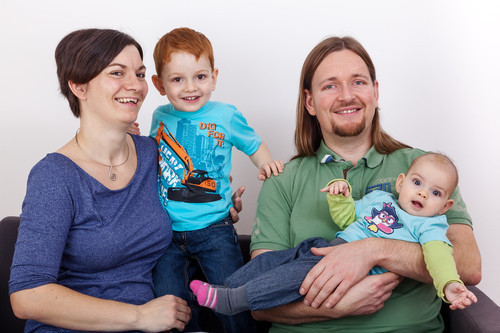 Eine vierköpfige Familie sitzt lächelnd auf einem Sofa. Links eine Frau mit dunklem Haar, daneben ein kleiner Junge im hellblauen T-Shirt, gehalten von der Frau. Rechts ein Mann mit langem Haar hält ein Baby im Arm, beide ebenfalls lächelnd. Der Junge trägt ein bunt bedrucktes T-Shirt mit einem Motiv. Der Kontext deutet auf Themen wie Arbeitsschutz im Familienhaushalt hin.