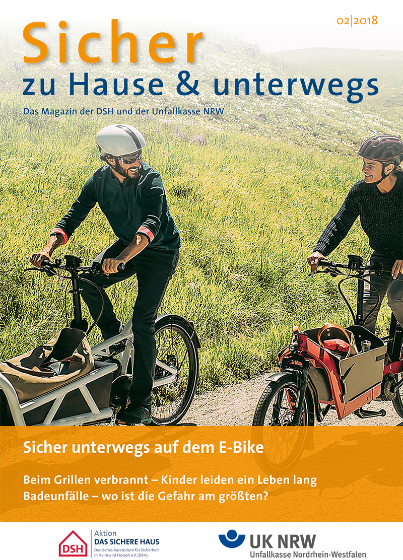 Zwei Erwachsene, ein Mann und eine Frau, fahren lächelnd auf E-Bikes auf einem gepflasterten Weg durch eine grüne Landschaft. Beide tragen Fahrradhelme. Im Hintergrund sind sanfte Hügel und Bäume zu sehen. Die Bildunterschrift thematisiert die Sicherheit bei E-Bike-Touren und verweist auf Themen wie die Prävention von Unfällen im Kontext des Arbeitsschutzes. Oben steht der Titel des Magazins 'Sicher zu Hause & unterwegs'. Logos der Unfallkasse NRW und der Aktion DAS SICHERE HAUS sind unten abgebildet.