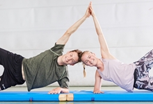Ein Mann und eine Frau liegen auf einer Gymnastikmatte und sind in der Seitenplanke-Position. Sie stützten sich auf dem rechten Arm ab und verlaufen diagonal zur Matte. Ihre nach oben gerichteten Hände berühren sich. Die Umgebung wirkt wie ein Fitnessstudio. Das Bild passt zur Prävention von Gesundheitsproblemen und Arbeitsunfällen durch körperliche Aktivität.
