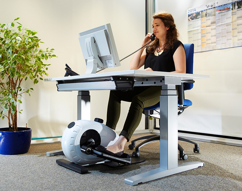 Eine Frau sitzt an einem ergonomischen Schreibtisch und telefoniert, während sie einen Beintrainer unter dem Tisch benutzt. Der Arbeitsplatz ist mit einem großen Monitor ausgestattet. Im Hintergrund sind eine pinnwandartige Wand und eine Pflanze zu sehen. Der Fokus liegt auf der Förderung von Bewegung am Arbeitsplatz zur Prävention von gesundheitlichen Problemen im Kontext von Arbeitsschutzmaßnahmen.