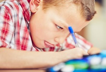 Ein junger Junge sitzt an einem Tisch und schreibt fokussiert mit einem blauen Kugelschreiber auf ein Blatt Papier. Er trägt ein rot-weiß kariertes Hemd und hat eine ernsthafte Miene. Im Vordergrund sind unscharfe Farben zu sehen, möglicherweise von einem Etui oder einer Federtasche. Das Bild könnte im Kontext der Unfallkasse NRW das Thema Sicherheit und Gesundheit in Schulen ansprechen, indem es die Bedeutung von geeigneten Arbeitsbedingungen und Materialien beim Schreiben und Lernen hervorhebt.